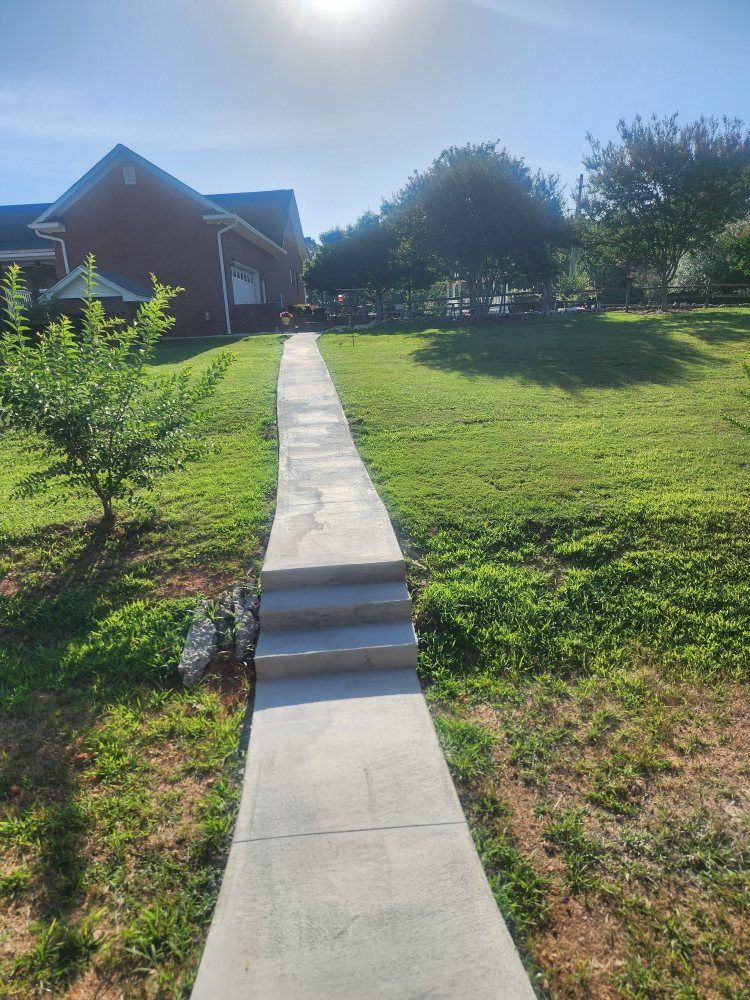 Concrete walkway with steps leading to a brick house across a sunny green lawn.