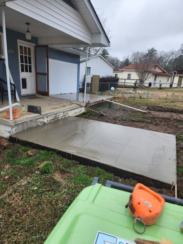 Concrete patio and walkway beside a house, with a green and orange wheelbarrow in the foreground.