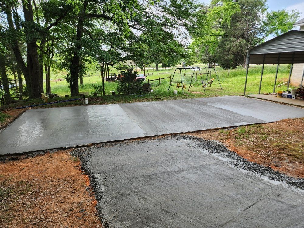 Fresh concrete driveway and patio extension beside a covered carport in a wooded yard