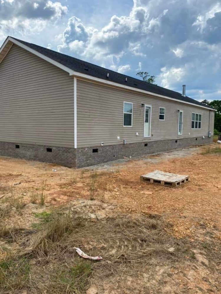 Single-story beige house with dark roof on a dirt lot under a cloudy sky