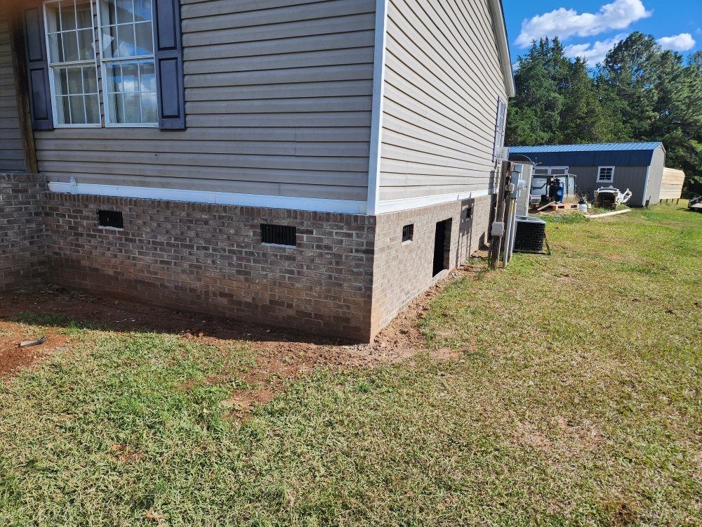 Side of a beige house with brick foundation, small windows, and grassy yard beside a detached shed.