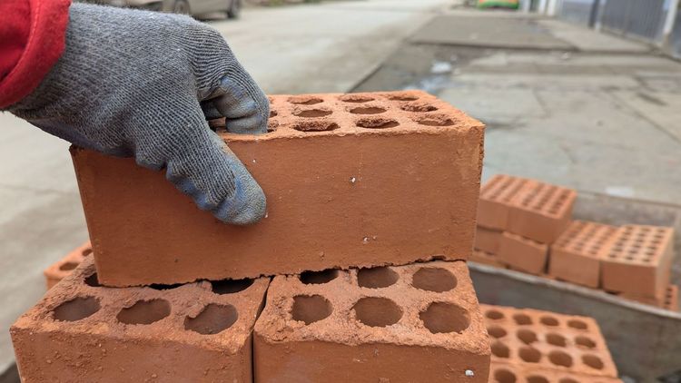 Gloved hand placing a red perforated brick on a stack of bricks at a construction site