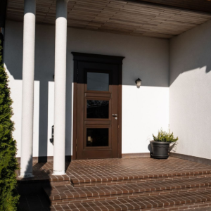 Front porch with white columns, wooden door, brick steps, and a potted plant in sunlight