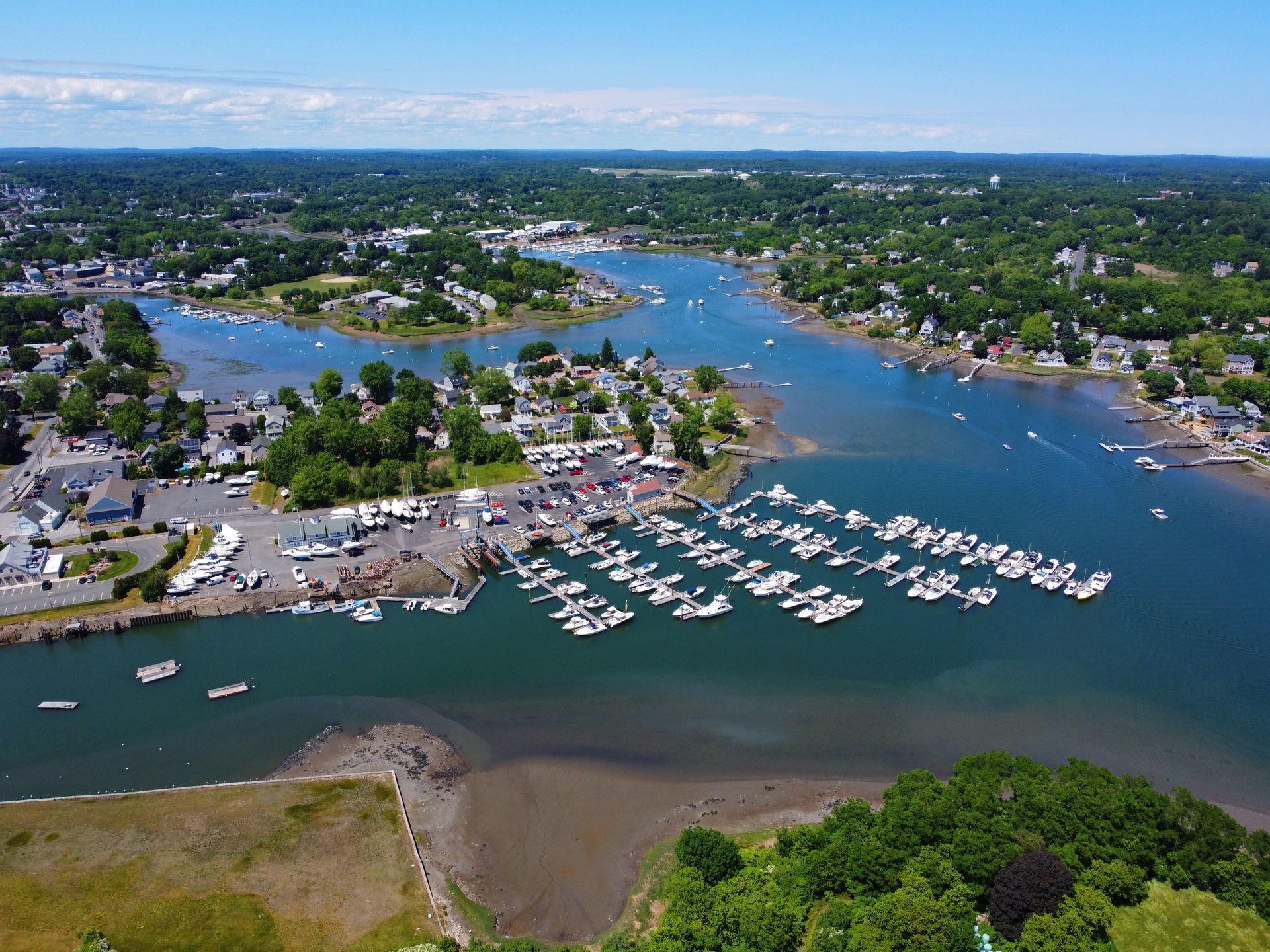Aerial view of a river harbor with many boats, shoreline homes, and green trees under a blue sky