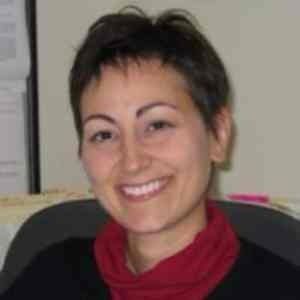 A person with short dark hair, wearing a red top under a black layer, smiles while looking toward the camera.