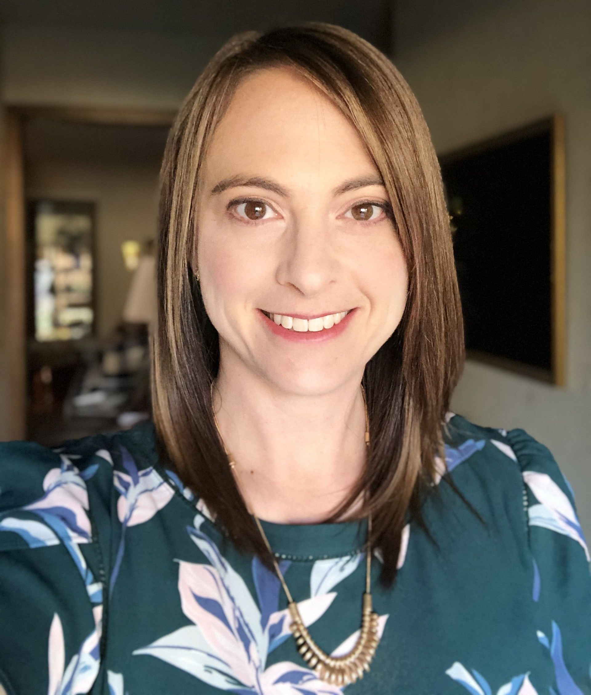 A smiling person with shoulder-length brown hair, wearing a dark green floral-patterned top.