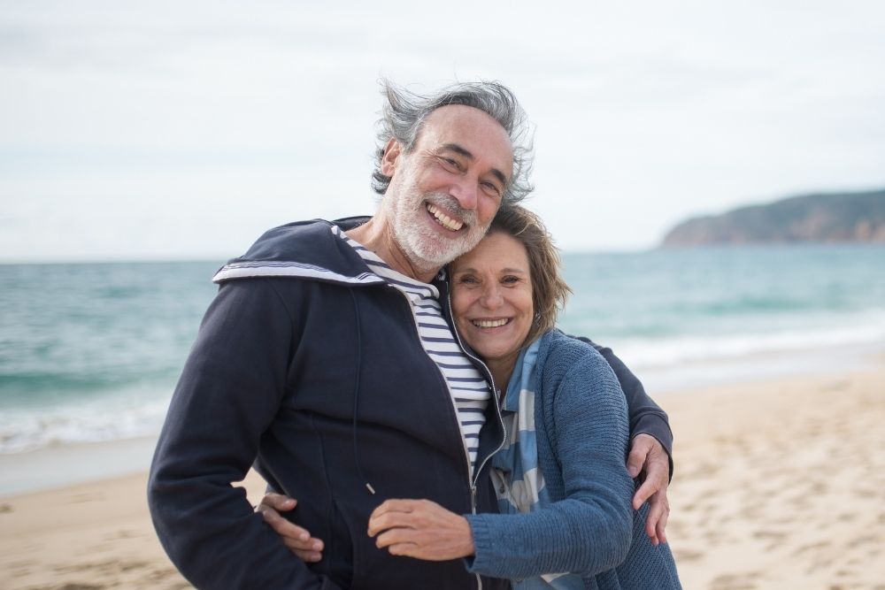 Man and woman embracing on a beach, both smiling. The man wears a dark jacket, the woman a blue cardigan. Ocean in background.