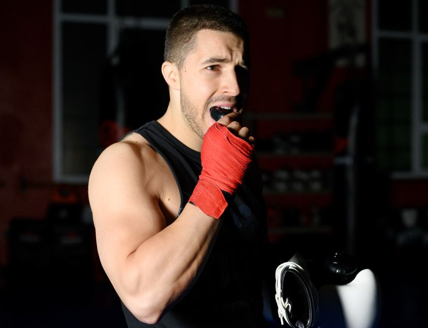 Boxer with mouthguard, wrapped hand near mouth, holding glove in gym.