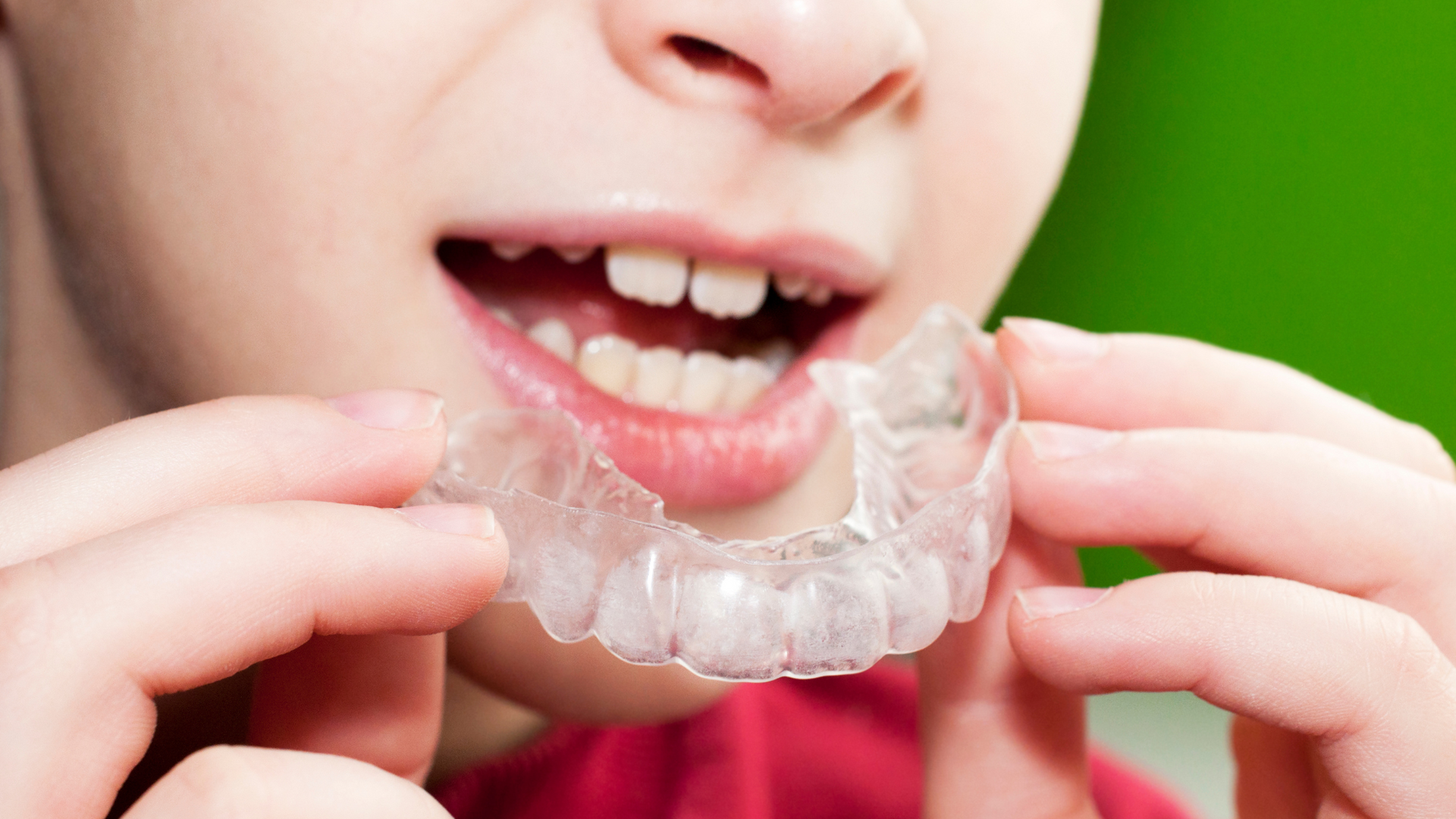 Person holding clear dental aligner near open mouth, green backdrop.