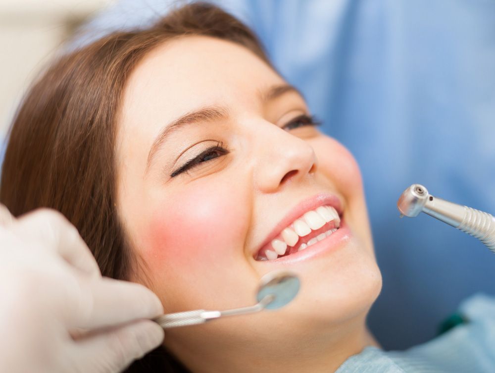 Woman at dentist, smiling during examination. Tools visible, blue setting.