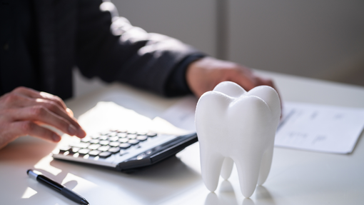 A person calculating dental costs with a calculator and a tooth model on a white desk.