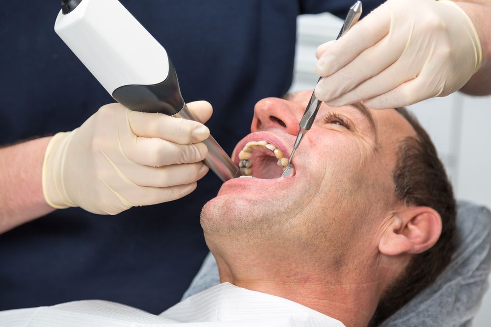 Dentist examining a patient's open mouth with dental tools. White gloves, blue shirt, neutral setting.