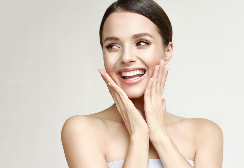 Woman with fair skin smiles widely, touching cheeks, against a light gray background.