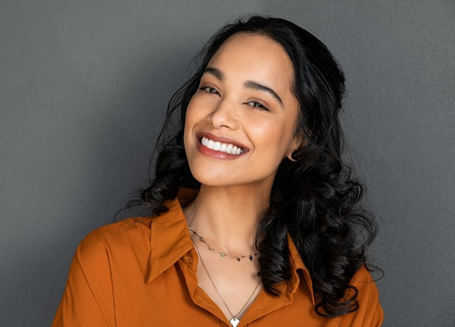 Woman with dark curly hair smiles at the camera, wearing an orange shirt and necklace, against a grey background.