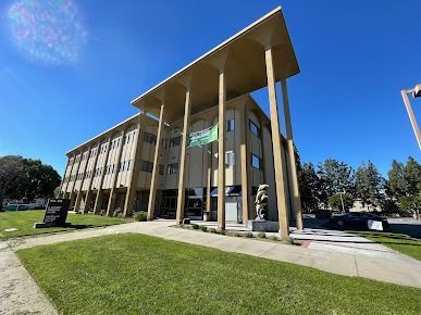Three-story beige building with tall pillars and large overhang, a green lawn and blue sky.