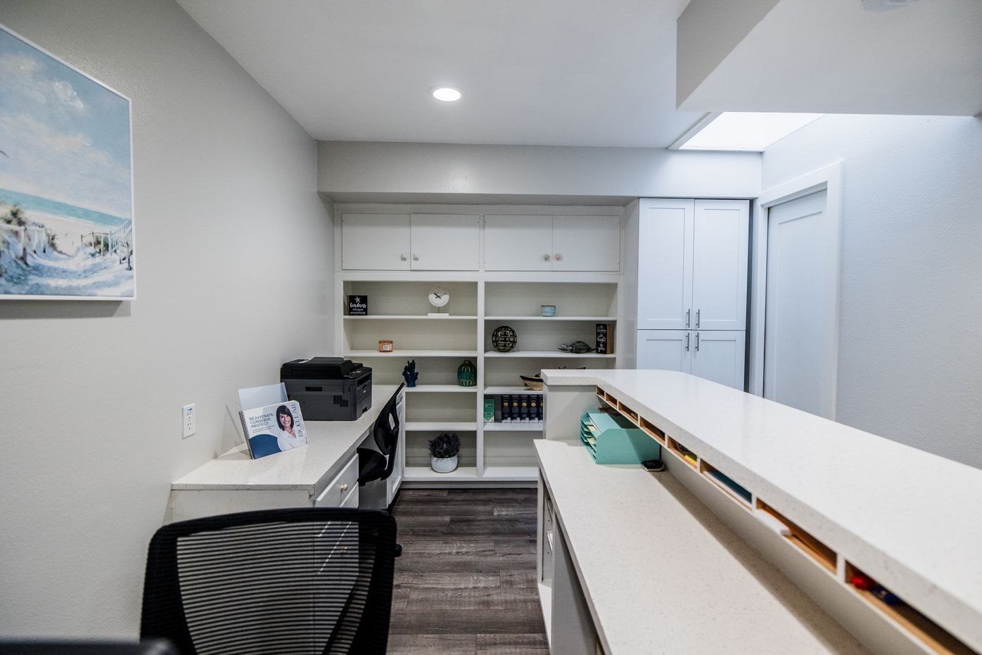 Office with white cabinets, shelving, and countertop, with a printer and artwork on the wall.
