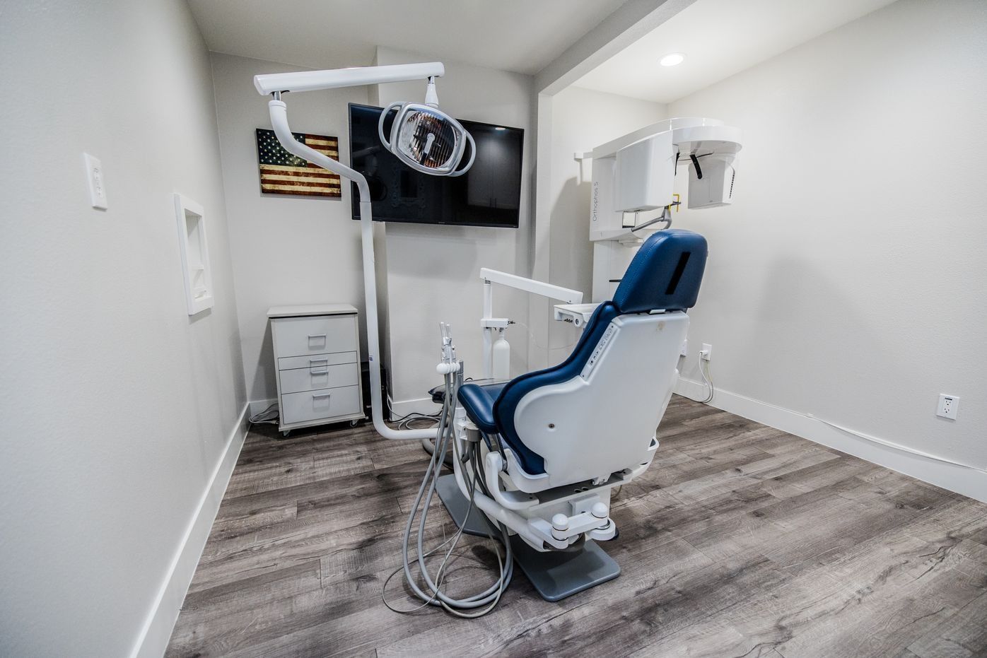 Dental operatory with chair, light, and X-ray equipment; a television hangs on the wall.