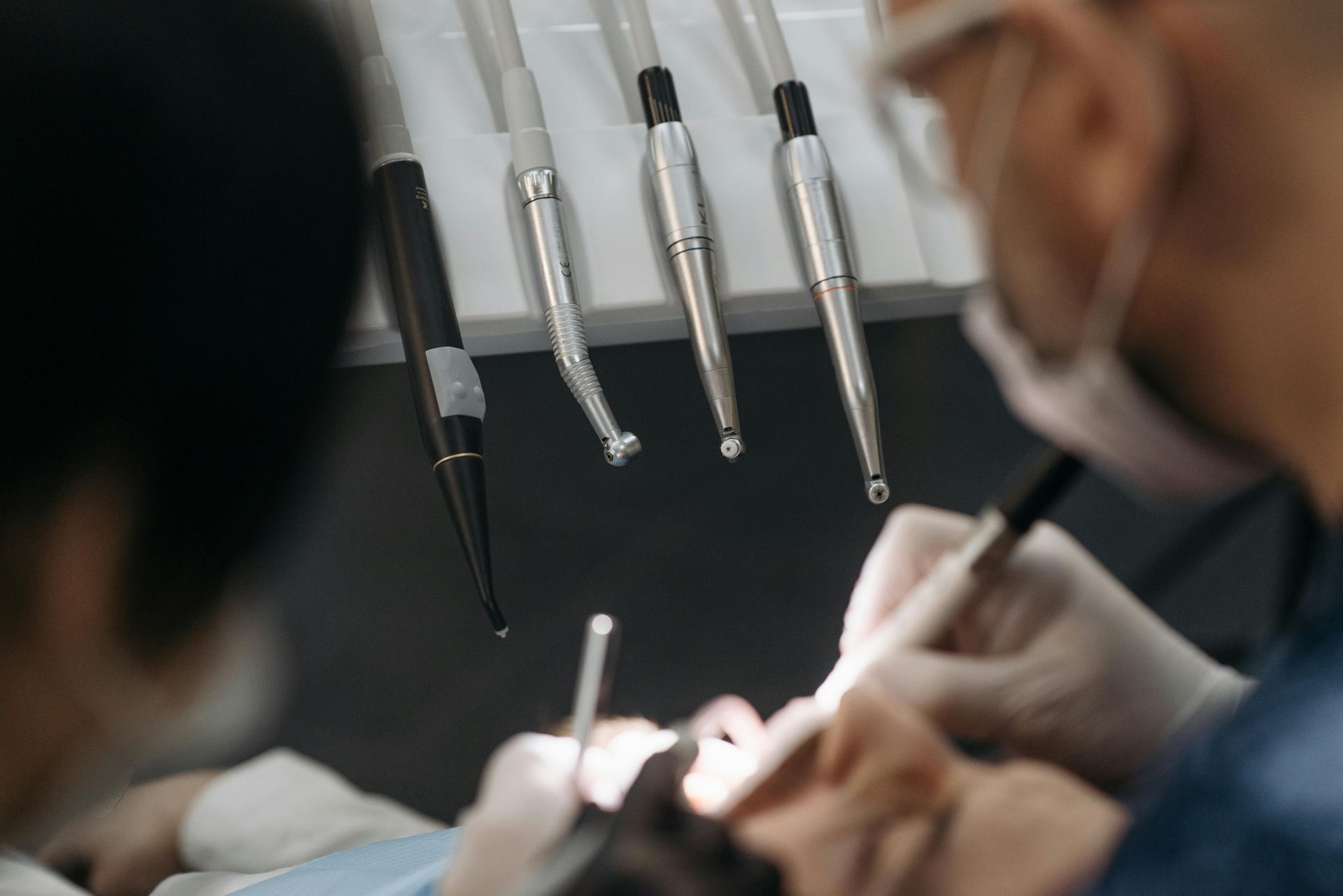 Woman in dental chair receiving examination, mouth open. Dentist holds tools, another assists.