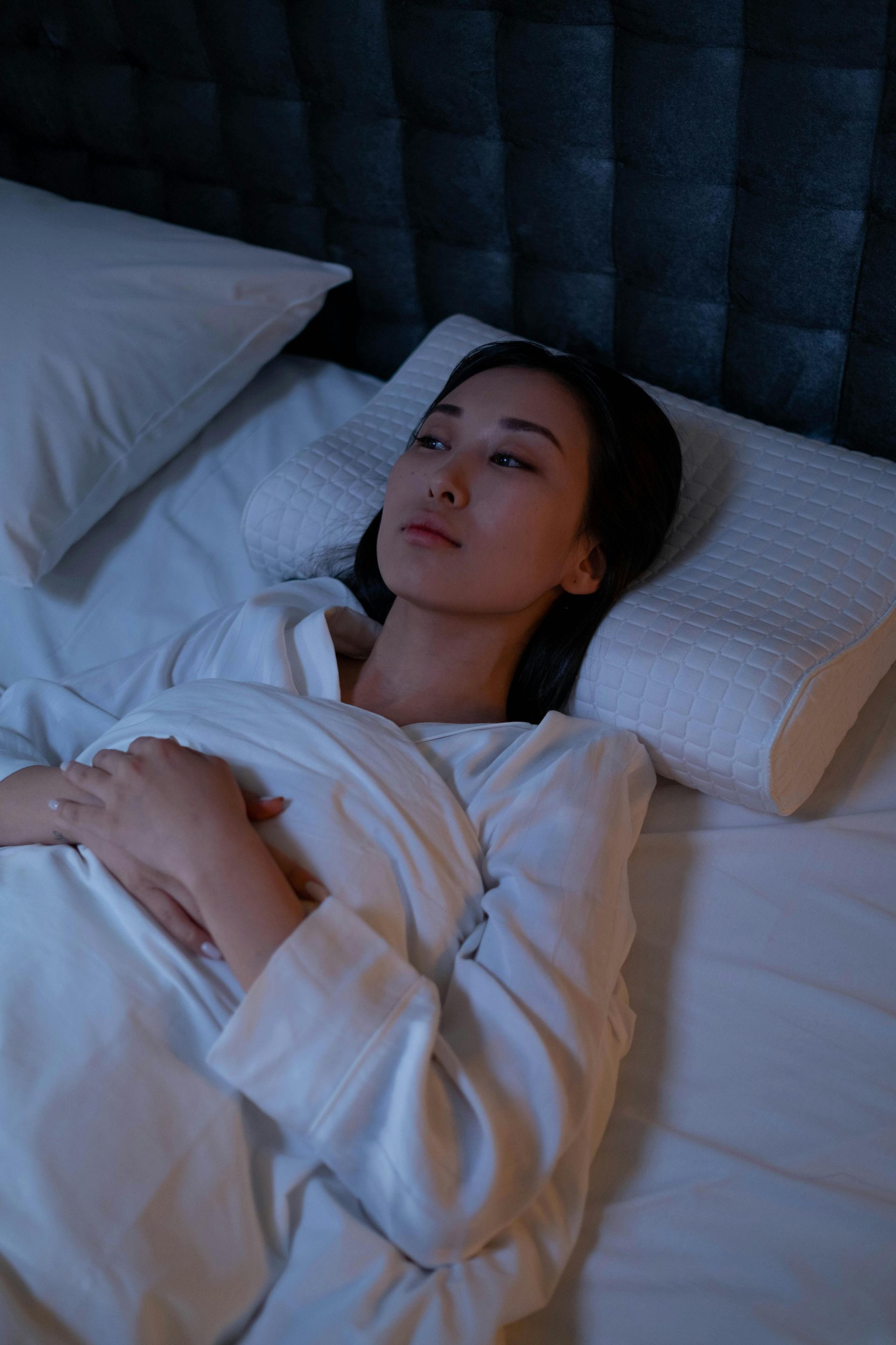 Woman in a white robe lying awake in bed, looking thoughtful.