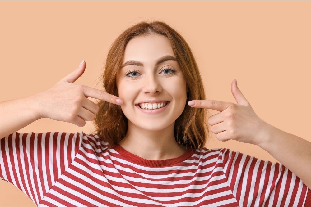 Woman with red-striped shirt smiling, pointing at her teeth with both index fingers. Light orange background.