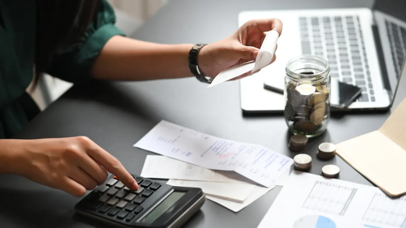 Person calculating expenses with receipts, calculator, and jar of coins on desk.