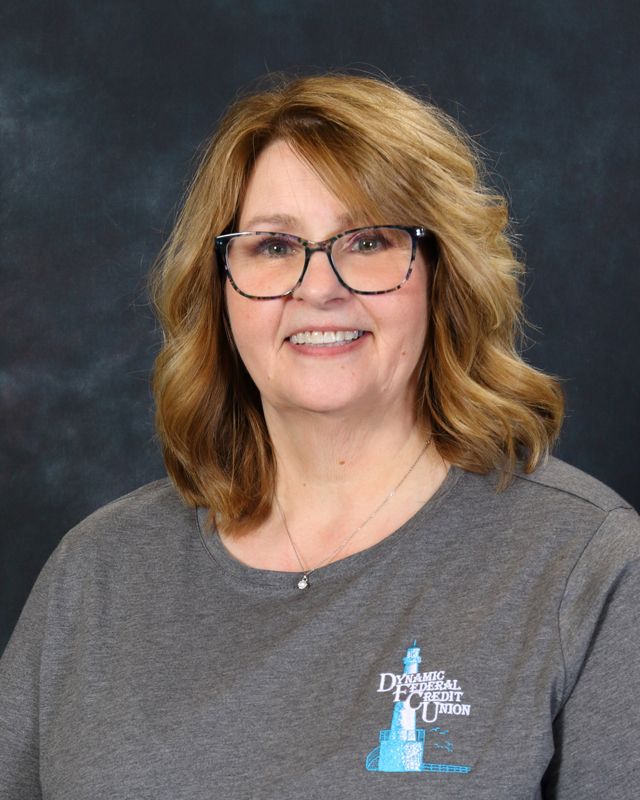 Woman with short blonde hair smiling, wearing a light blue button-up shirt with logo, against a teal background.