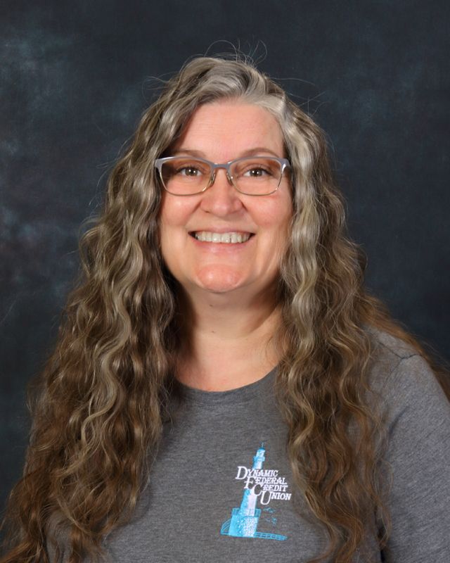 Woman with glasses smiling, wearing a grey button-down shirt, standing in front of a teal background.