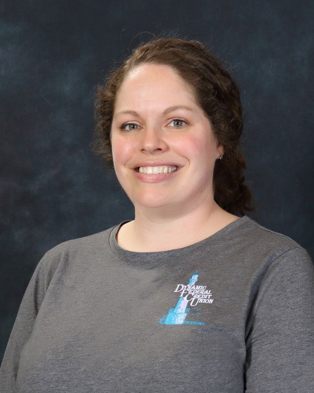 Woman with brown hair and smiling wearing a gray button-up shirt with an emblem, against a blue background.