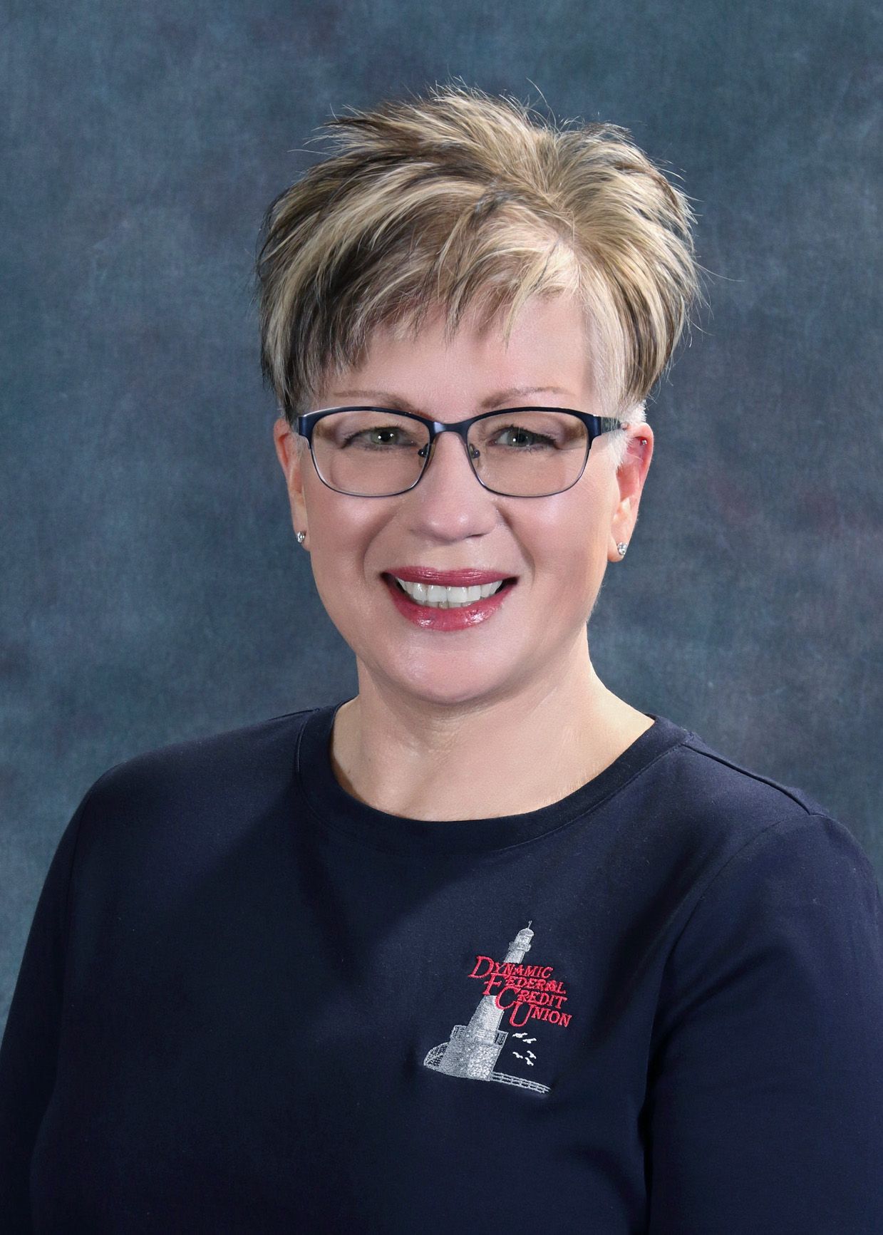 Woman with glasses, short blonde hair, wearing a navy shirt smiling at the camera.