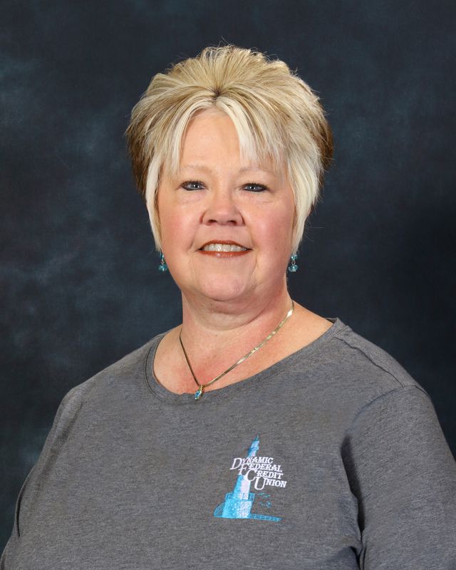 Woman with short blonde hair smiles, wearing a light blue button-up shirt against a blue background.