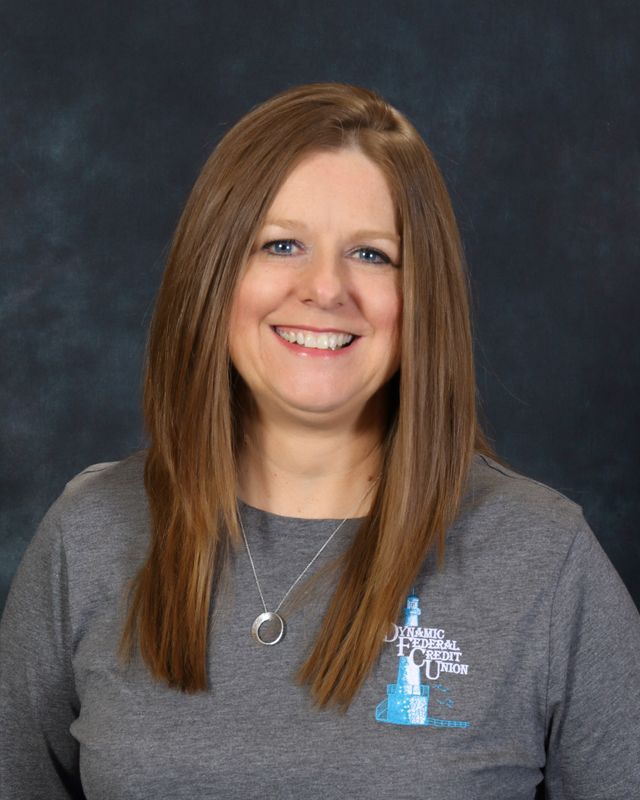 Woman with long brown hair, smiling, wearing a gray button-down shirt with logo on teal background.