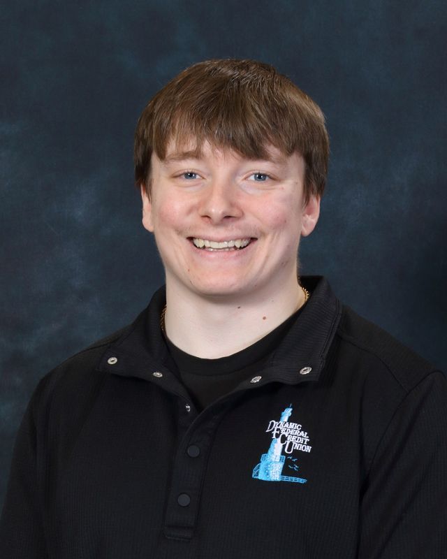 Smiling young man wearing a grey button-down shirt with a logo, standing against a blue background.