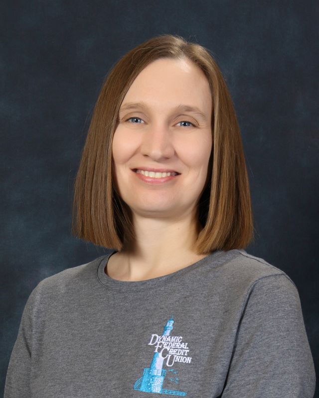 Woman with light brown hair, smiling, wearing a gray shirt with a logo on a blue background.