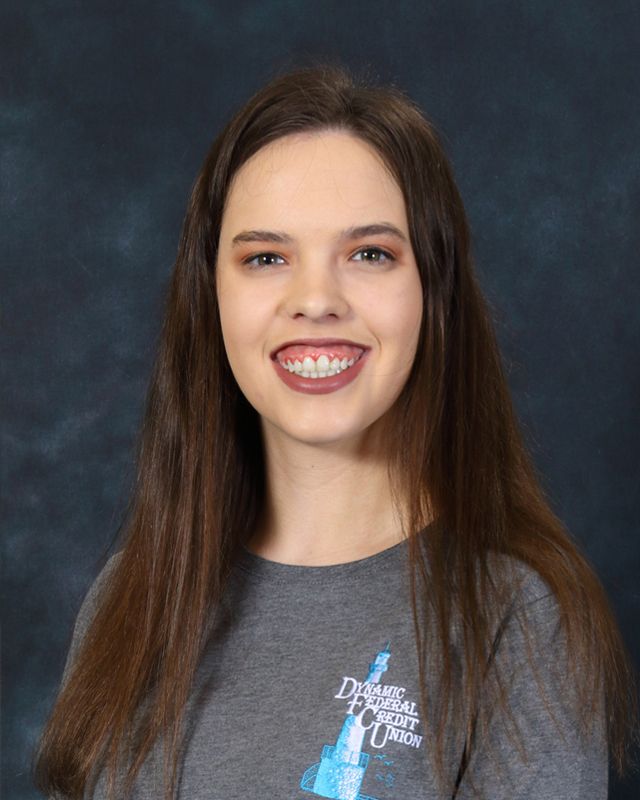 Woman with long brown hair, smiling, wearing a gray button-down shirt and pearl earrings, against a blue background.