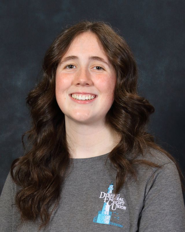 Woman with long brown hair, smiling, wearing a gray button-down shirt and pearl earrings, against a blue background.