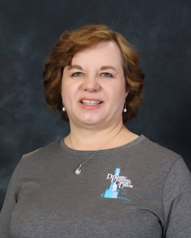 Woman with curly brown hair, smiling, wearing a gray shirt with a logo, against a blue background.