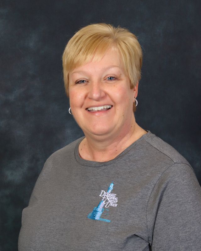 Woman with short blonde hair smiling, wearing a light blue button-up shirt with logo, against a teal background.