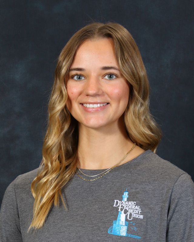 Woman with curly brown hair, smiling, wearing a gray shirt with a logo, against a blue background.