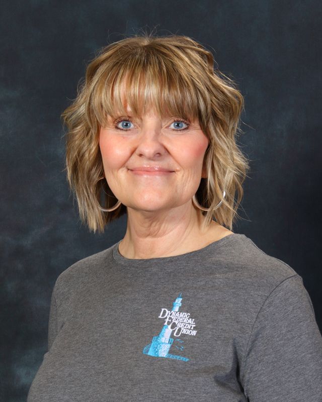 Woman with long brown hair, smiling, wearing a gray button-down shirt with logo on teal background.