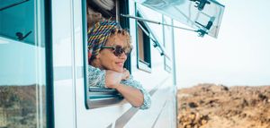 Woman in sunglasses and headscarf leans out of RV window, smiling. Desert landscape.