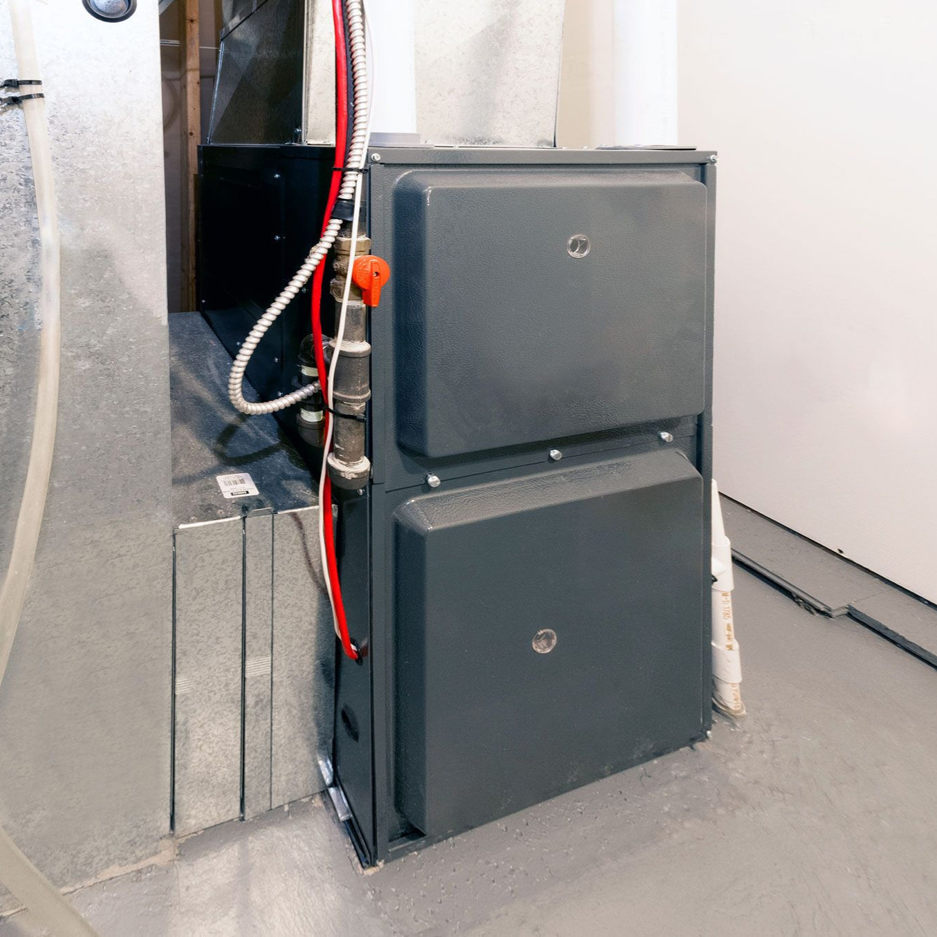 A gray gas furnace stands indoors with a red gas shut-off valve and wiring visible on its side.