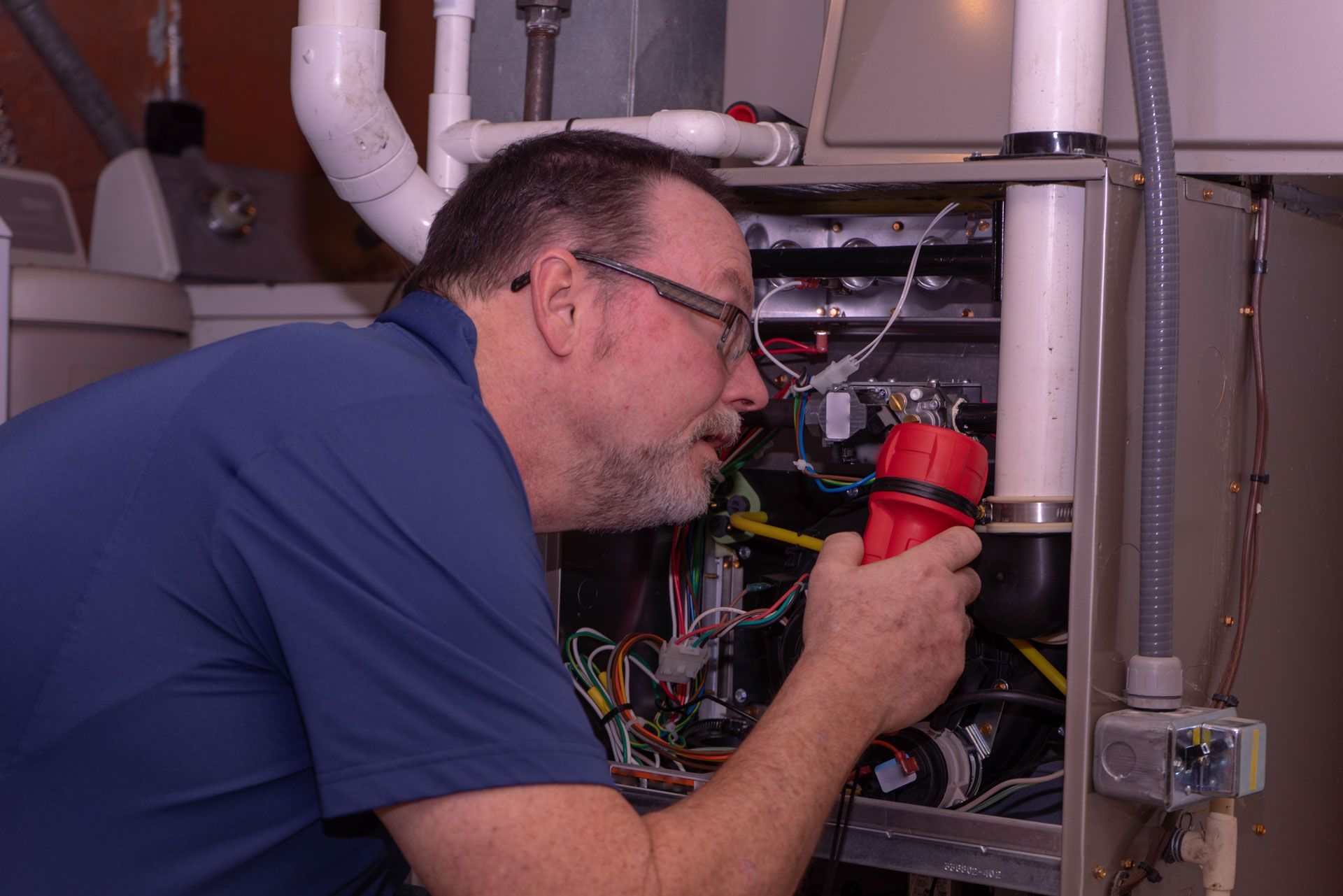 A technician in a blue shirt uses a red flashlight to inspect the internal wiring of an open HVAC furnace.