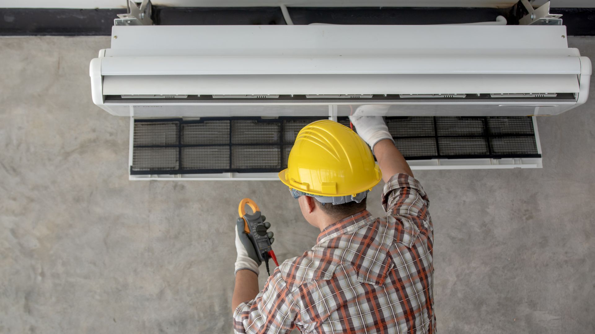A technician in a yellow hard hat and plaid shirt uses a multimeter to inspect an open wall-mounted air conditioner.