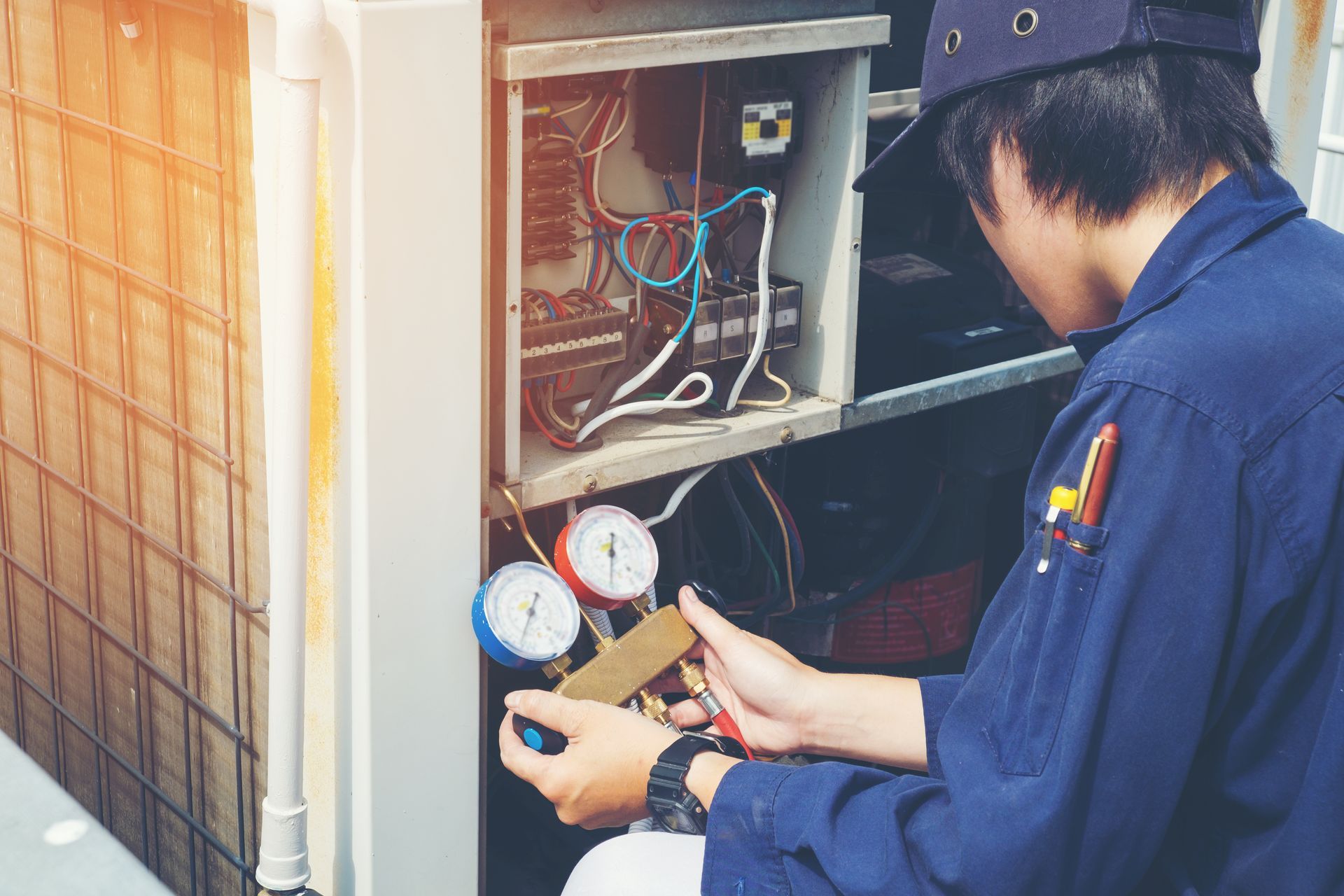 A technician in a blue uniform inspecting an open outdoor HVAC unit while holding a manifold gauge set.