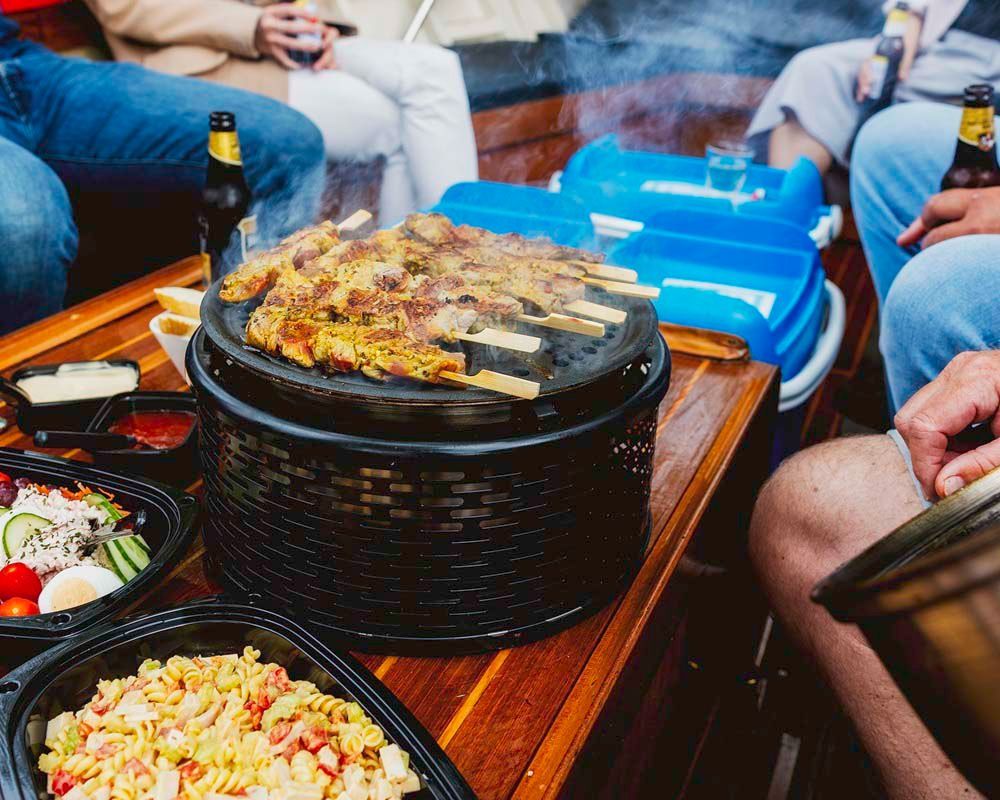 Mensen hadden zich verzameld rond een tafel met een sissende grill, schalen met eten en bierflesjes op een boot.