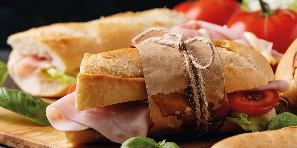Een sandwich, omwikkeld met keukentouw, gevuld met ham en sla, met daarnaast tomaten en groene bladgroenten op een boot.