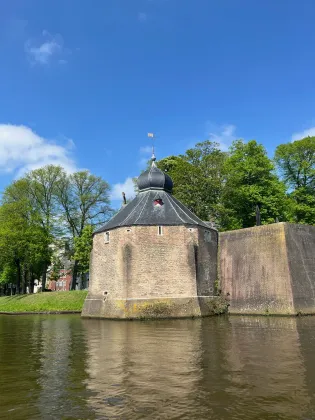 Historische stenen toren naast een gracht, met groene bomen en een blauwe lucht die weerspiegeld wordt in het kalme water.