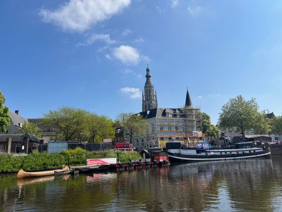 Een tafereel van een kanaal met boten en een kerktoren die weerspiegeld worden in het kalme water onder een blauwe hemel.