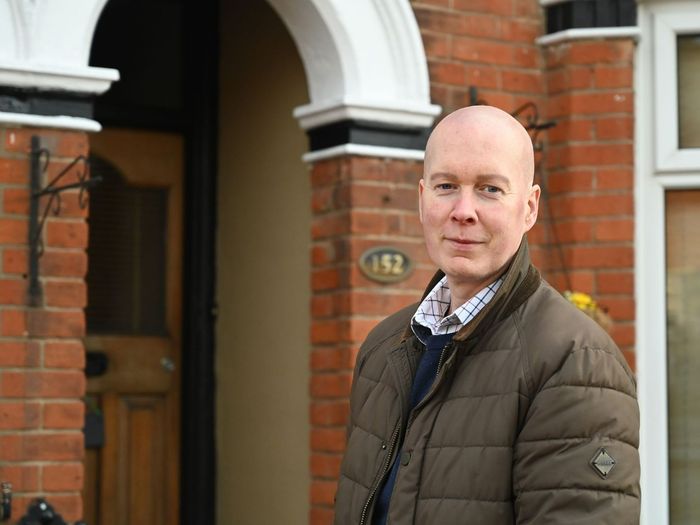 A bald man in a brown jacket is standing in front of a brick house.