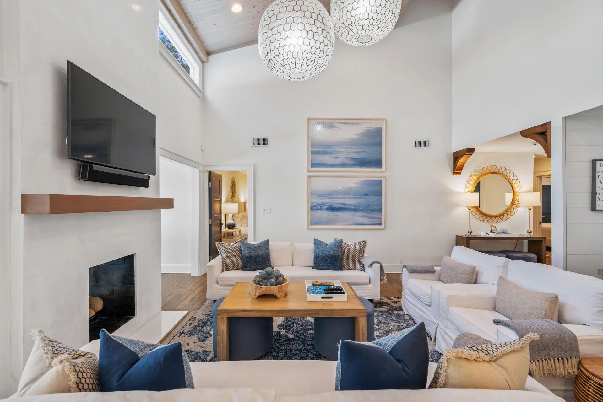 Bright, airy living room with white sofas, blue accents, fireplace, wall TV, and ocean artwork.